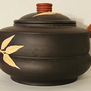 May include: A black ceramic teapot with a bamboo-style handle and spout, and a lid with a small, red knob. The teapot is decorated with gold-colored leaf accents. The handle and spout are a reddish-brown color.