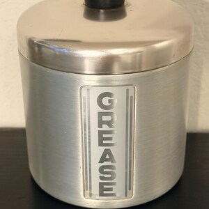 May include: A cylindrical, brushed metal grease container with a lid and a black knob. The word "GREASE" is vertically displayed in a rectangular, frosted window on the front. The container is designed for kitchen use.
