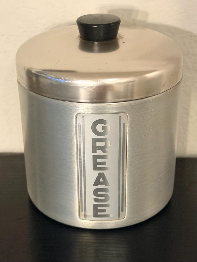 May include: A cylindrical, brushed metal grease container with a lid and a black knob. The word "GREASE" is vertically displayed in a rectangular, frosted window on the front. The container is designed for kitchen use.
