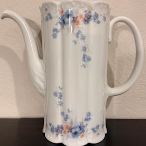 May include: A white porcelain coffee pot with a scalloped rim and base, featuring a delicate floral pattern of light blue and soft orange blossoms. The vessel includes a curved handle, a long spout, and a matching lid with a small handle.