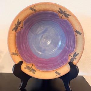 May include: A round ceramic bowl with a wide, textured tan rim featuring black dragonfly illustrations. The interior of the bowl has a smooth, wavy-edged basin transitioning from a deep purple outer ring to a light blue center.