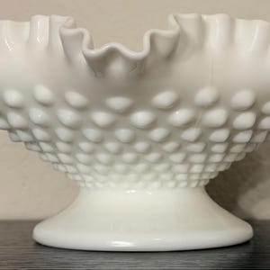 Puede incluir: Un cuenco de vidrio de leche blanco con borde con volantes. El cuenco tiene una base de pedestal y una superficie texturizada con pequeñas protuberancias espaciadas uniformemente. El borde con volantes añade un toque decorativo.