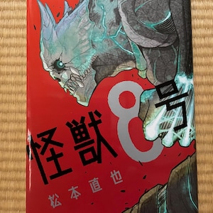 Könnte beinhalten: Ein Manga-Band mit rotem Cover, das eine detaillierte Illustration eines Monsters mit leuchtendem türkisblauem Design zeigt. Der Titel ist in japanischen Schriftzeichen, mit der Nummer "1" oben links. Das Logo "Jump Comics+" befindet sich unten.