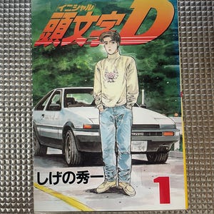 May include: A manga cover featuring a young man in a beige turtleneck and blue jeans, standing in front of a white car. The cover has Japanese text and the number 1 in red. The background is green.