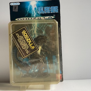 May include: A plastic blister pack containing a black Godzilla action figure. The packaging features a blue and white background with lightning bolts and the text "Real Hero Series" and "Godzilla".