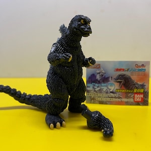 Peut inclure: Figurine articulée de Godzilla noire avec un bras détaché, sur une surface jaune. La figurine a des griffes et des nageoires dorsales jaunes. Une petite boîte avec du texte japonais est derrière.