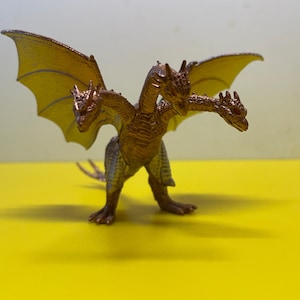 Puede incluir: Una figura de dragón de tres cabezas de color bronce, con alas extendidas. El dragón tiene un cuerpo y alas detallados y texturizados, de pie sobre una superficie amarilla. Las alas son de un tono amarillo más claro, contrastando con el cuerpo.
