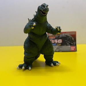 Puede incluir: Figura de acción verde de Godzilla con expresión sonriente, de pie sobre una superficie amarilla. La figura tiene escamas y garras detalladas. Una caja con texto japonés es visible en el fondo. La figura mide unos 15 cm de altura.