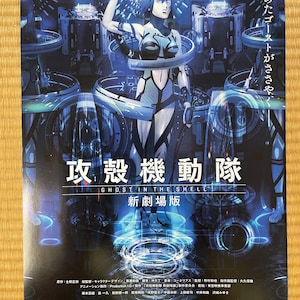 以下が含まれることがあります： 映画『攻殻機動隊 新劇場版』のポスター。青と黒のサイバネティックなスーツを着た中央の人物が、青く光る機械の中に立っている。ポスターには「攻殻機動隊」の文字と2015年初夏ROADSHOWの記載がある。
