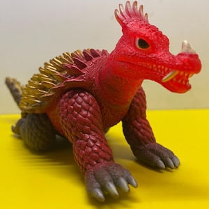 Puede incluir: Monstruo de juguete de plástico rojo y dorado con una cresta puntiaguda y dientes afilados.