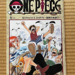May include: A One Piece manga volume 1, Romance Dawn, featuring a colorful cover illustration of three characters on a wooden ship deck. The title One Piece is printed in large black letters at the top, with the author name Eiichiro Oda below.