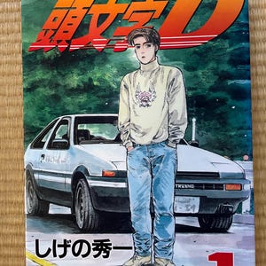 Könnte beinhalten: Ein Cover eines japanischen Mangas mit einem jungen Mann in einem weißen Pullover und blauen Jeans, der neben einem weißen Auto steht. Der Titel des Mangas ist "Initial D" und die Nummer des Bandes ist "1".