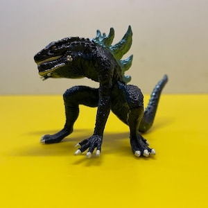 Könnte beinhalten: Eine schwarze und teal Godzilla-Figur mit strukturiertem Körper und Rückenflossen. Das Spielzeug ist auf einer gelben Oberfläche platziert und zeigt seine detaillierten Merkmale und seine reptilienartige Form. Die Figur hat weiße Krallen und einen langen Schwanz.