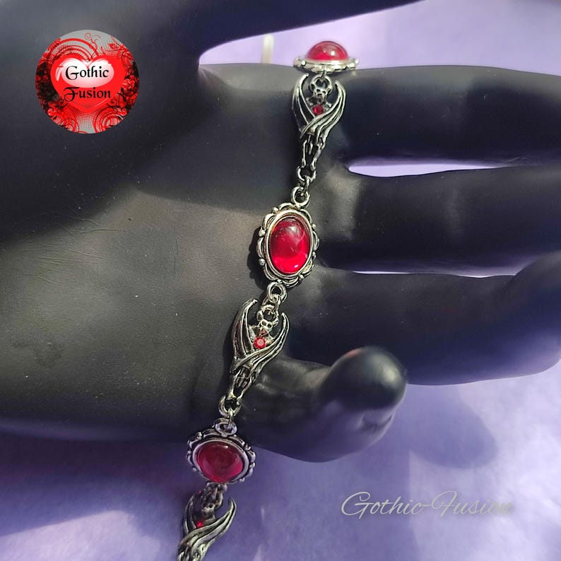 May include: A silver bracelet with red gemstone accents and bat wing charms. The bracelet is on a black surface.