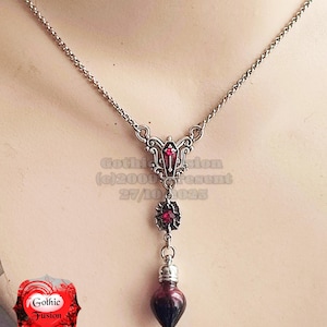 May include: Silver-toned pendant necklace with a gothic design. The necklace features a decorative top piece with a red gem, a smaller gem pendant, and a teardrop-shaped vial filled with dark red liquid. The text "Gothic Fusion" is visible.