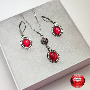 May include: A silver necklace and earrings set with red gemstone pendants. The necklace features a small diamond-shaped charm with a red gemstone in the centre. The earrings are drop earrings with a red gemstone in a silver oval setting.