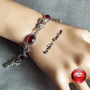 May include: A silver bracelet with red gemstone accents. The bracelet features a delicate, ornate design with swirling patterns. The text "Gothic-Fusion" is written on the wrist.