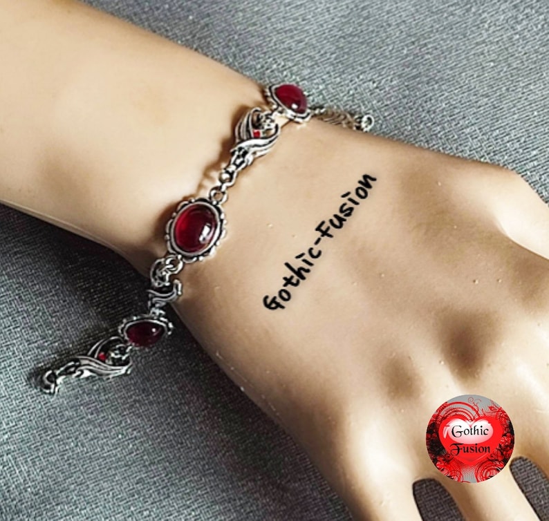 May include: A silver bracelet with red gemstone accents. The bracelet features a delicate, ornate design with swirling patterns. The text "Gothic-Fusion" is written on the wrist.