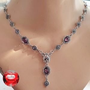 May include: A silver necklace with a pendant featuring a large purple gemstone. The necklace has a delicate chain with smaller purple gemstones.