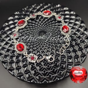 May include: A silver bracelet with red gemstone accents. The bracelet has a clasp closure and is displayed on a black background.