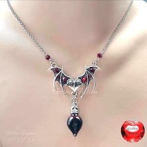 May include: A silver necklace with a bat wing pendant and a red glass vial. The pendant is adorned with red crystals. The necklace is on a white background.