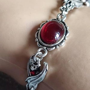 May include: Silver bracelet with red gemstone accents and a bat charm. The bracelet has a delicate chain and ornate detailing.