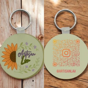 May include: A circular keyring with a silver loop. One face shows a sunflower and the text "Artisan Lav" in purple and green. The other side has a QR code and the Instagram handle @ARTISANLAV in orange.