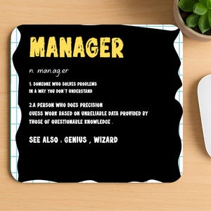 May include: A black mouse mat with a definition of "MANAGER" in yellow and white text. The definition includes two points describing a manager's role. The mouse mat is on a wooden desk next to a keyboard, mouse, and a small potted plant.