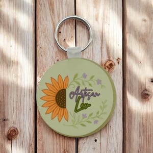 May include: A round keyring with a sage green background featuring a yellow sunflower on the left side and a green leafy vine wreath on the right. The text Artisan Lav is printed in the centre in a dark purple and green script font.