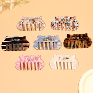 Puede incluir: Una colección de peines personalizados para el cabello en varios colores y patrones. Cada peine presenta un nombre o iniciales únicos en letras doradas. Los peines tienen forma de cabeza de gato y están hechos de un material brillante.