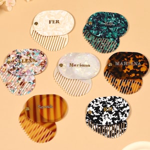 May include: A selection of personalised combs in different colours and patterns. Each comb has a matching oval case with a name or initials in gold lettering. The combs are in shades of white, green, brown, and black.