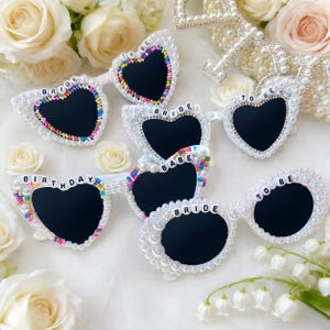 May include: White heart-shaped sunglasses with black lenses, decorated with pearls and colourful beads. Some sunglasses have text that reads "BRIDE", "TO BE", "BIRTHDAY", and "BABE". These sunglasses are a wedding or party accessory.