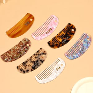 May include: Seven personalized combs in various colors and patterns, including tortoiseshell, pink marble, and white. Each comb has a name or initials engraved on it, such as "Julia" and "Sophie". The combs are arranged on a light orange surface.