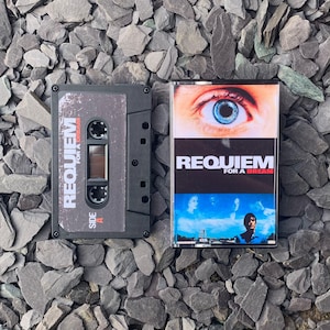 May include: Two cassette tapes with the title "Requiem for a Dream." One tape is black with white text, the other has a movie poster design with a blue eye and the film title. The tapes are on a bed of gray rocks.