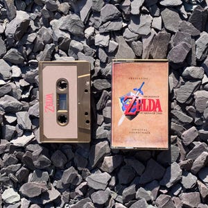 May include: A gold-coloured cassette tape and case featuring the text "Zelda" and "The Legend of Zelda: Ocarina of Time Original Soundtrack." The cassette tape is a vintage music format.