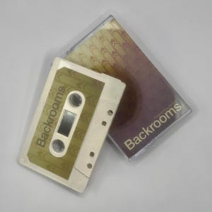 Peut inclure: Une cassette audio blanche avec une étiquette dorée portant le texte Backrooms et un motif de flèches répétitives, accompagnée d'un boîtier en plastique transparent contenant un insert au dégradé violet et doré avec le texte Backrooms.