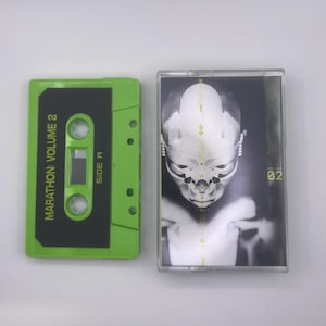 May include: A green cassette tape labelled "MARATHON: VOLUME 2" beside a cassette case with a black and white graphic of a face. The cassette tape is green with black accents. The cassette case has the number "02" on it.