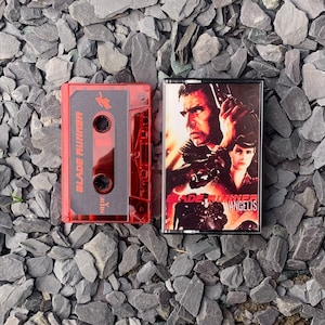 May include: A red cassette tape labeled "Blade Runner" next to a cassette case with movie art. The tape is translucent, revealing the internal components. The case features a movie poster with actors and the film title.