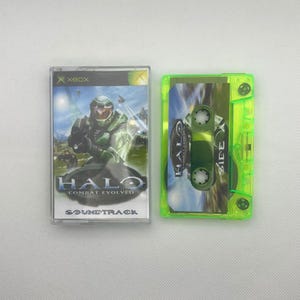 May include: A neon green cassette tape and case featuring the Halo: Combat Evolved soundtrack. The case displays the game's cover art with a green-armoured character. The cassette tape is visible inside the case.