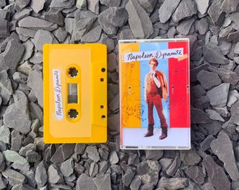 May include: Two yellow cassette tapes. One tape has the text "Napoleon Dynamite" on a white label. The other tape has the same text on a white label and a colorful image of a man in a brown suit.