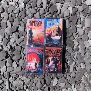 Puede incluir: Se muestran cuatro novelas de ciencia ficción de Dan Simmons. Las portadas presentan ilustraciones vibrantes con títulos como "Hyperion" y "The Fall of Hyperion". Los libros están dispuestos sobre un lecho de piedras grises.