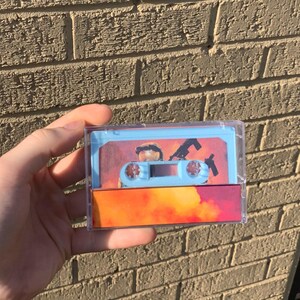 May include: A light blue cassette tape with a clear plastic case. The cassette tape has a picture of a person in a military uniform holding a gun. The background of the picture is a fiery orange and yellow explosion.