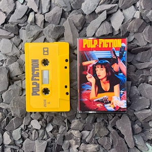 Puede incluir: Una cinta de cassette amarilla y una caja de cassette con el título de la película "Pulp Fiction". La cinta de cassette es amarilla con el título de la película impreso verticalmente. La caja de cassette tiene una imagen de la película de una mujer.