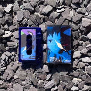 May include: Two blue cassette tapes. One has a colourful abstract design, the other displays a blue-toned anime illustration with the text "NEON GENESIS EVANGELION". The tapes are set against a background of dark grey rocks.