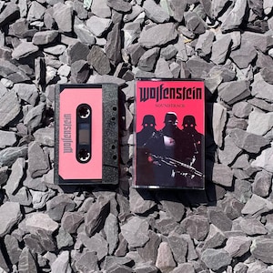 Op de afbeelding: Twee cassettes met het woord "Wolfenstein". Eén is roze met witte tekst, de andere heeft een rode cover met de woorden "Wolfenstein Soundtrack" en een afbeelding van soldaten. De cassettes liggen op een achtergrond van grijze stenen.