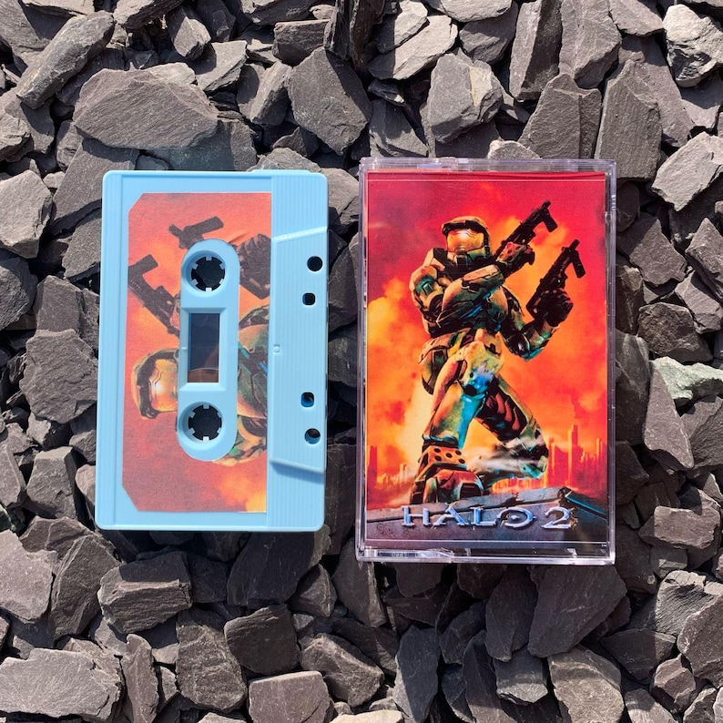 May include: Two blue cassette tapes with a red and orange image of a video game character holding a gun. The text "HALO 2" is printed on the tape.