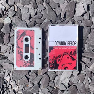 May include: Two white cassette tapes with red and black artwork. One tape features a red graphic design, while the other displays the text "COWBOY BEBOP" in white. The tapes are set against a background of dark gray rocks.