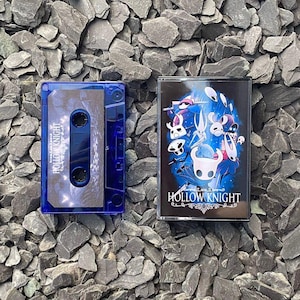 Peut inclure: Une cassette audio bleue et son boîtier avec le texte "Hollow Knight" et des illustrations de personnages. Le boîtier a un fond bleu foncé avec des motifs blancs et bleus. La cassette est bleu transparent.