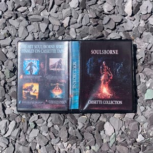 Puede incluir: Una caja de cinta de cassette negra con el texto "SOULSBORNE CASSETTE COLLECTION". La caja está abierta, revelando los títulos de los juegos "DARK SOULS" y "ELDEN RING". La caja está sobre un fondo de rocas grises.