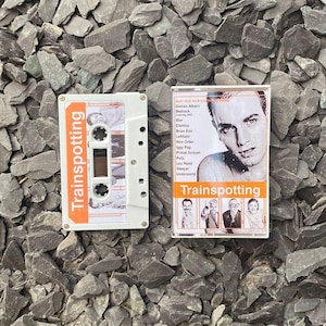 以下が含まれることがあります： 「Trainspotting」と書かれたオレンジ色のラベルが付いた白いカセットテープとそのケース。ケースには男性の写真と曲のリストが掲載されています。カセットテープは灰色の岩の上にあります。